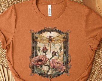 May include: A rust-colored t-shirt with a vintage-style illustration of a dragonfly and flowers. The illustration is framed in a dark brown border with gold accents.