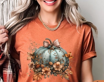 May include: A teal blue pumpkin with a brown bow and fall leaves on a rust colored t-shirt. The shirt is labeled "Bella Canvas Heather Autumn".