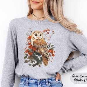 May include: A gray long-sleeve shirt with a white barn owl perched on a branch with red berries and green leaves. The shirt is Bella Canvas 3501 Athletic Heather.