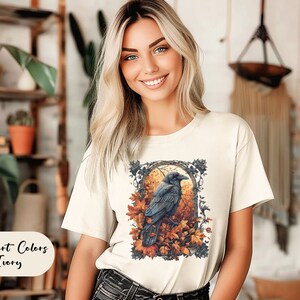 May include: Ivory-colored t-shirt featuring a detailed illustration of a raven perched amidst autumn leaves. The design is framed within an ornate border. The text "Comfort Colors Ivory" is displayed on the lower left.