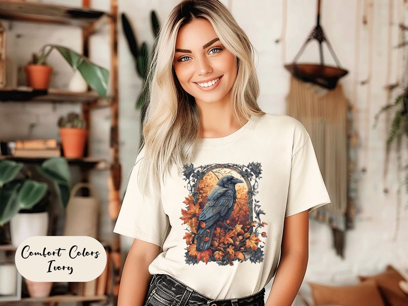 May include: Ivory-colored t-shirt featuring a detailed illustration of a raven perched amidst autumn leaves. The design is framed within an ornate border. The text "Comfort Colors Ivory" is displayed on the lower left.