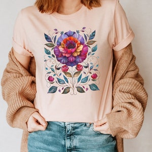 May include: A pink t-shirt with a colorful floral design. The design features a large flower with pink, purple, and orange petals, surrounded by green leaves and vines.