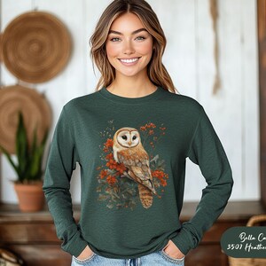 May include: A dark green long-sleeved shirt with a white-faced barn owl with orange and brown markings perched on a branch with red berries. The owl is surrounded by green leaves and the text "Bella Canvas 3501 Heather Forest" is in the bottom right corner.