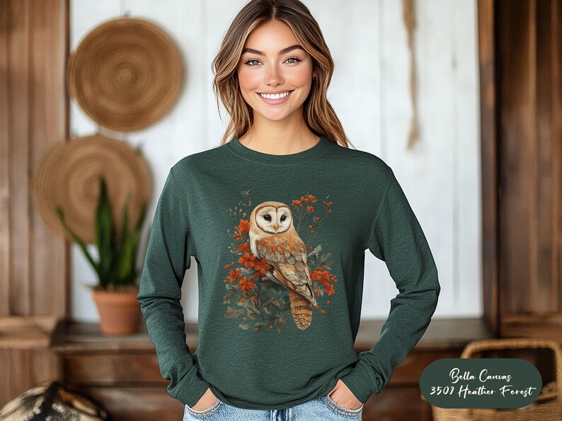 May include: A dark green long-sleeved shirt with a white-faced barn owl with orange and brown markings perched on a branch with red berries. The owl is surrounded by green leaves and the text "Bella Canvas 3501 Heather Forest" is in the bottom right corner.