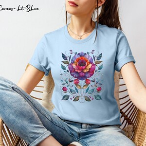 May include: A light blue t-shirt with a floral design featuring a large purple, red, and orange flower with green leaves. The text "Bella Canvas - Lt Blue" is visible at the top of the image.