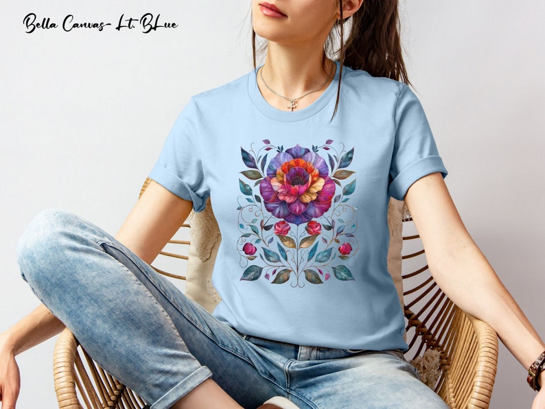 May include: A light blue t-shirt with a floral design featuring a large purple, red, and orange flower with green leaves. The text "Bella Canvas - Lt Blue" is visible at the top of the image.