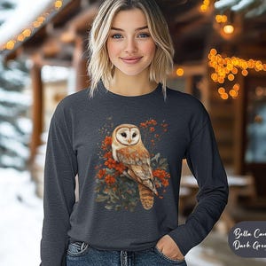 May include: A dark grey heather Bella + Canvas 3507 long-sleeve shirt with a white barn owl perched on a branch with red berries. The owl has a white face and brown and white feathers.