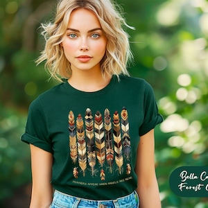 May include: A woman wearing a forest green t-shirt with a design of colorful feathers. The shirt has the text "FEATHERS APPEAR WHEN ANGELS ARE NEAR". The text is in gold lettering. The background is a blurred out green.