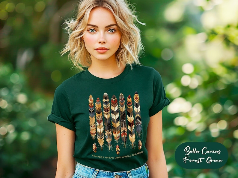 May include: A woman wearing a forest green t-shirt with a design of colorful feathers. The shirt has the text "FEATHERS APPEAR WHEN ANGELS ARE NEAR". The text is in gold lettering. The background is a blurred out green.