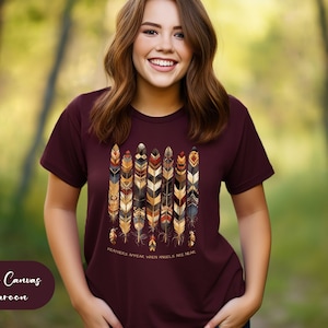 May include: A maroon Bella Canvas t-shirt with a graphic of colorful feathers and the text "FEATHERS APPEAR WHEN ANGELS ARE NEAR".