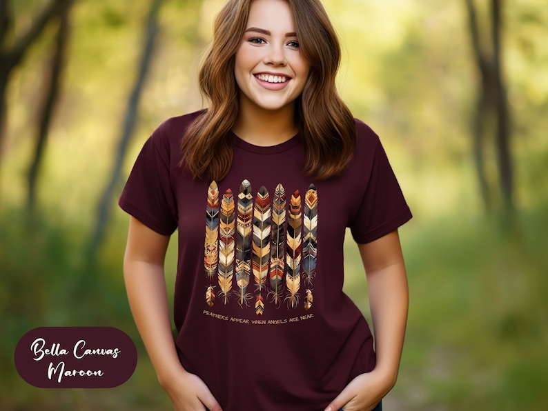May include: A maroon Bella Canvas t-shirt with a graphic of colorful feathers and the text "FEATHERS APPEAR WHEN ANGELS ARE NEAR".