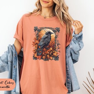 May include: Terracotta-colored t-shirt featuring a detailed illustration of a raven perched amidst autumn leaves. The artwork is framed within an ornate border. The text "Comfort Colors Terracotta" is displayed at the bottom left.