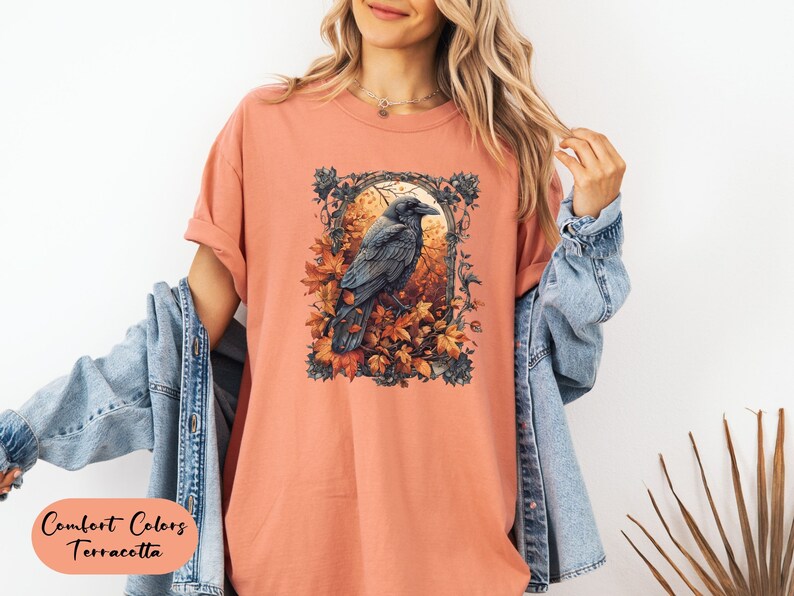 May include: Terracotta-colored t-shirt featuring a detailed illustration of a raven perched amidst autumn leaves. The artwork is framed within an ornate border. The text "Comfort Colors Terracotta" is displayed at the bottom left.