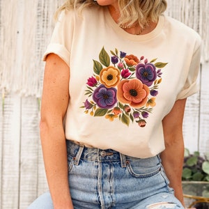 May include: A cream colored t-shirt with a floral print featuring purple, orange, yellow, and green flowers.