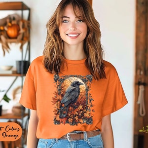 May include: Burnt orange t-shirt featuring a detailed illustration of a raven perched amidst autumn leaves and a full moon. The shirt's text reads "Comfort Colors Burnt Orange". The model is wearing a brown belt and blue jeans.