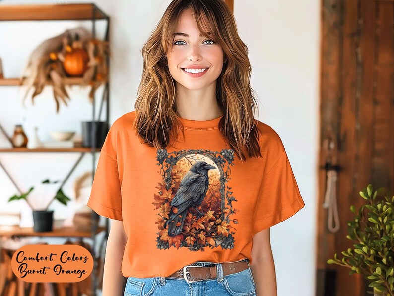 May include: Burnt orange t-shirt featuring a detailed illustration of a raven perched amidst autumn leaves and a full moon. The shirt's text reads "Comfort Colors Burnt Orange". The model is wearing a brown belt and blue jeans.