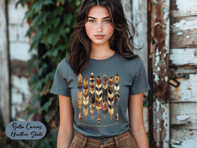 May include: A heather slate gray t-shirt with a graphic of stylized feathers in shades of brown, gold, and red. The text "FEATHERS APPEAR WHEN ANGELS ARE NEAR" is printed below the graphic.