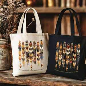 May include: Two tote bags, one cream and one black, with a feather design. The cream bag has a white handle, and the black bag has a black handle. The text "Feathers appear when angels are near" is printed on the bottom of each bag.