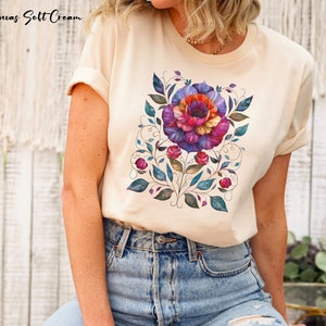 May include: A cream colored t-shirt with a floral design featuring a large purple, pink, and orange flower with green leaves and vines. The design is centered on the shirt.