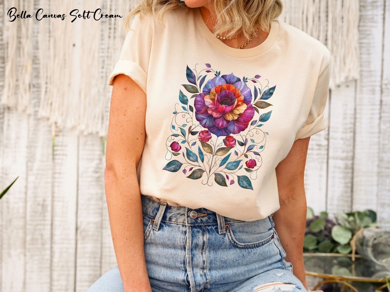 May include: A cream colored t-shirt with a floral design featuring a large purple, pink, and orange flower with green leaves and vines. The design is centered on the shirt.