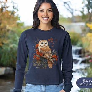 May include: A navy blue long-sleeved sweatshirt with a white barn owl perched on a branch with red berries. The owl is surrounded by green leaves and the text "Bella Canvas 3501 Heather Navy" is in the bottom right corner.