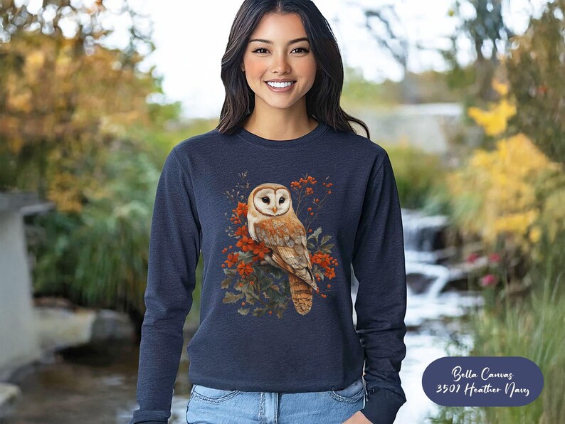 May include: A navy blue long-sleeved sweatshirt with a white barn owl perched on a branch with red berries. The owl is surrounded by green leaves and the text "Bella Canvas 3501 Heather Navy" is in the bottom right corner.