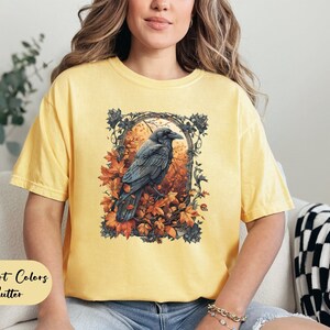 May include: Yellow Comfort Colors t-shirt with a graphic of a raven perched in an autumn scene. The raven is black, surrounded by orange and brown leaves. The text "Comfort Colors Butter" is at the bottom of the shirt.
