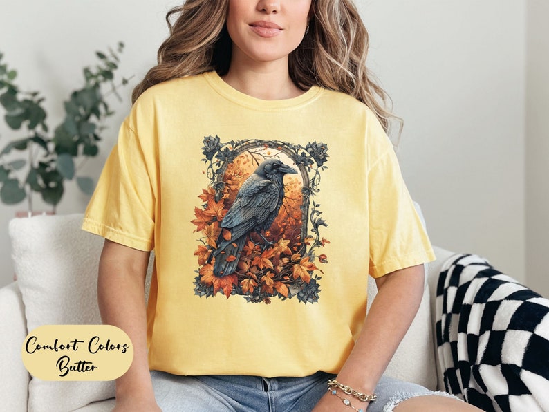 May include: Yellow Comfort Colors t-shirt with a graphic of a raven perched in an autumn scene. The raven is black, surrounded by orange and brown leaves. The text "Comfort Colors Butter" is at the bottom of the shirt.
