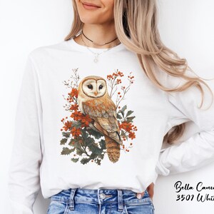 May include: White long-sleeve shirt with a brown and white barn owl perched on a branch with red berries and green leaves.