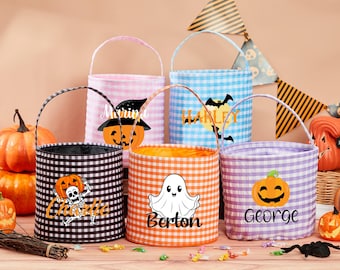May include: A selection of Halloween treat bags in gingham fabric, in black, pink, blue, orange, and purple. Each bag has a Halloween graphic and a name. Pumpkins and decorations are in the background.