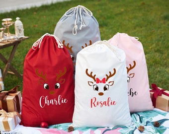 May include: Four drawstring gift bags in red, grey, white, and pink, each with a reindeer face design. The red bag says "Charlie" in white script, and the white bag says "Rosalie" in red script. The bags are on a blanket.