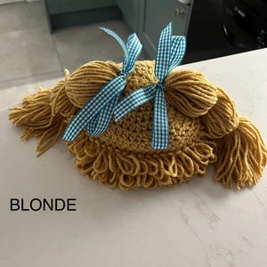 May include: A crocheted hat with two pigtails and blue and white gingham bows. The hat is a light brown color and the text "BLONDE" is visible below the hat.