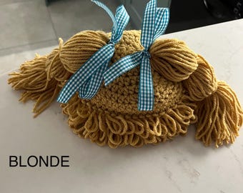 May include: A crocheted hat with two pigtails and blue and white gingham bows. The hat is a light brown color and the text "BLONDE" is visible below the hat.
