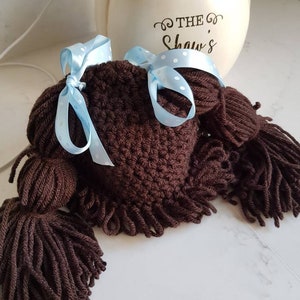 May include: A brown crocheted doll wig with two long brown pigtails and a blue ribbon with white polka dots.