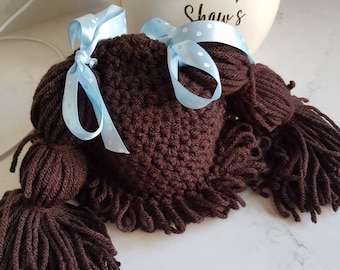 May include: A brown crocheted doll wig with two long brown pigtails and a blue ribbon with white polka dots.