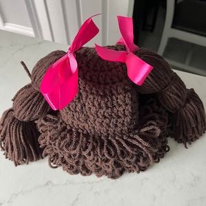 May include: Brown crocheted hat with two pigtails and pink bows. The hat is made of yarn and has a shaggy texture.