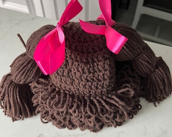 May include: Brown crocheted hat with two pigtails and pink bows. The hat is made of yarn and has a shaggy texture.