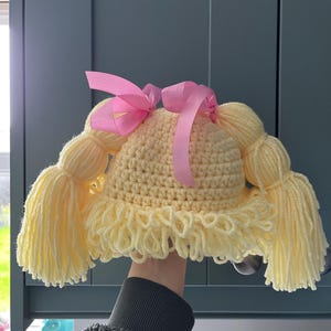May include: A pale yellow crocheted hat featuring two large yarn pigtails on the sides, a textured fringe along the bottom edge, and two bright pink satin ribbons tied at the top to secure the pigtails in place, creating a doll-like appearance.