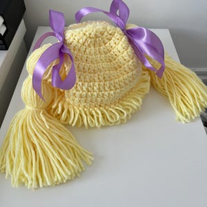 May include: A pale yellow crocheted hat featuring a textured, rounded crown and a fringe along the bottom edge. Two long, thick yarn pigtails extend from the sides, each secured with a smooth, shiny purple satin ribbon tied into a bow.
