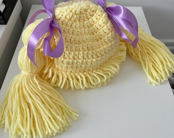 May include: A pale yellow crocheted hat featuring a textured, rounded crown and a fringe along the bottom edge. Two long, thick yarn pigtails extend from the sides, each secured with a smooth, shiny purple satin ribbon tied into a bow.