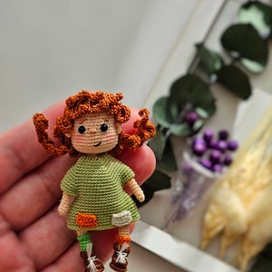 May include: A handmade crochet doll with red curly hair and a green dress. The doll has small brown shoes and patches on the legs. The doll is held in a hand, with a blurred background of greenery and flowers.