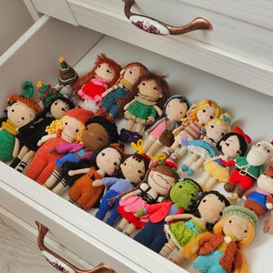 May include: A drawer filled with various handmade crochet dolls. The dolls have different skin tones, hair colors, and outfits, including dresses, overalls, and holiday-themed attire. The drawer is white with bronze-colored handles.