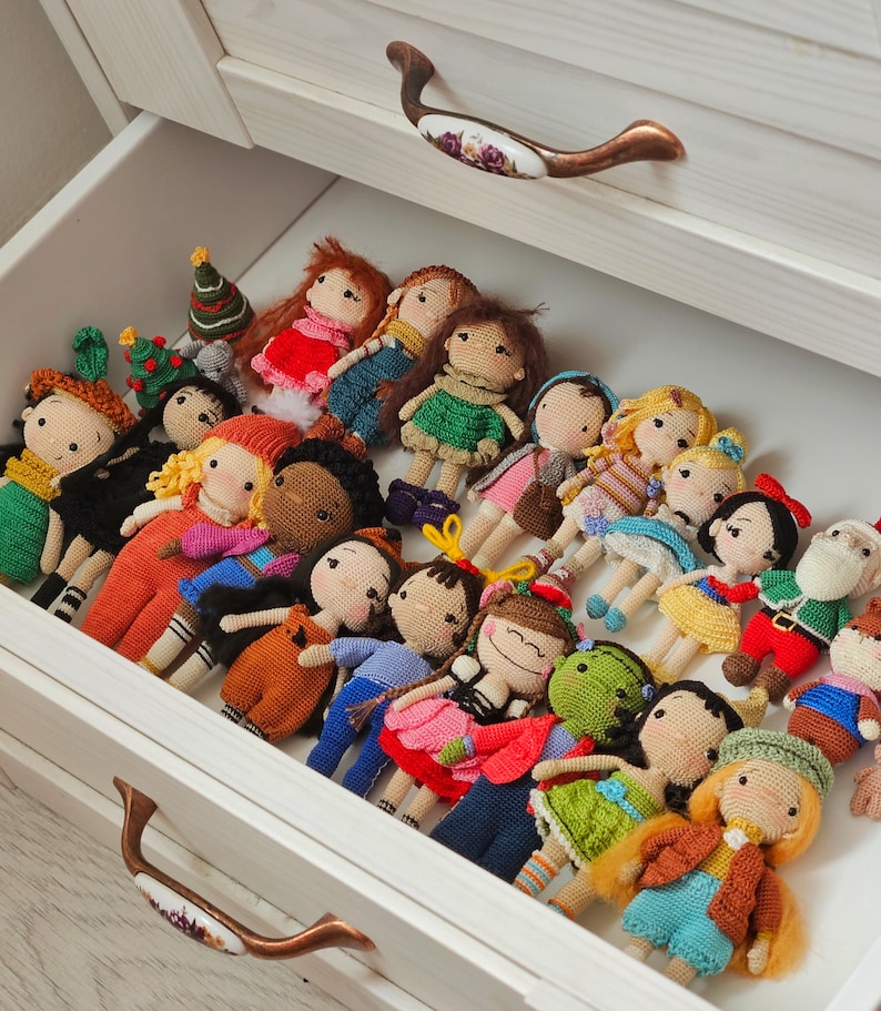 May include: A drawer filled with various handmade crochet dolls. The dolls have different skin tones, hair colors, and outfits, including dresses, overalls, and holiday-themed attire. The drawer is white with bronze-colored handles.