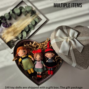 May include: Three handmade crochet dolls in a heart-shaped gift box with decorative paper. The dolls have red, orange, and brown hair, and wear dresses in green, pink, and black. The gift box is next to a heart-shaped box with a white ribbon.