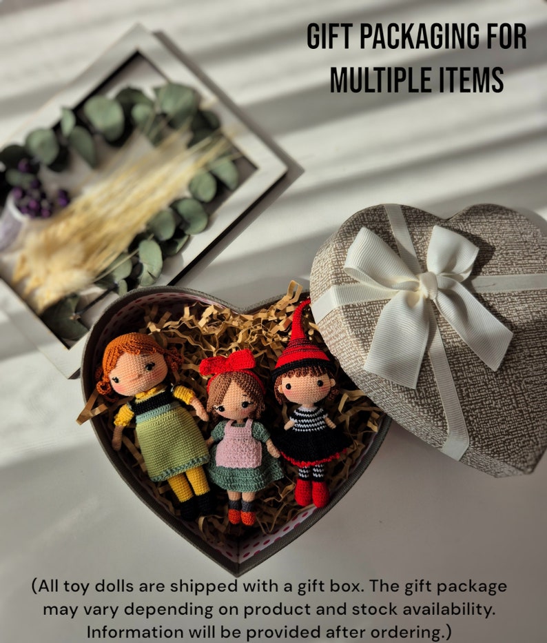 May include: Three handmade crochet dolls in a heart-shaped gift box with decorative paper. The dolls have red, orange, and brown hair, and wear dresses in green, pink, and black. The gift box is next to a heart-shaped box with a white ribbon.