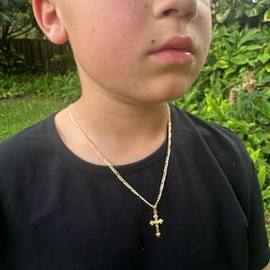 May include: A gold chain necklace with a small gold cross pendant. The necklace is worn by a person with a black shirt.
