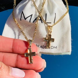 以下が含まれることがあります： 十字架のペンダントが付いたゴールドチェーンネックレス2本。チェーンは、「MALI」と印刷された白い布製のバッグの上に置かれています。