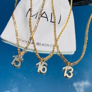 May include: Gold-toned necklaces with pendants featuring the numbers 13, 15, and 16. The necklaces have different chain styles, including a rope chain and a figaro chain. A white pouch with the word "MALI" is visible in the background.