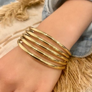 May include: A set of five gold-toned bangle bracelets stacked on a person's wrist.