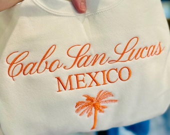 May include: White sweatshirt with orange embroidered text that reads "Cabo San Lucas MEXICO" and a palm tree graphic. The sweatshirt is held up by a hand.